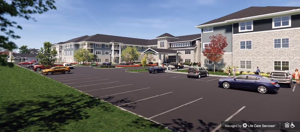 Exterior view of a large senior living facility building with multiple floors, a covered entrance, and a parking lot with several cars. There are a few people walking near the building, and trees and grass surround the area under a clear blue sky.
