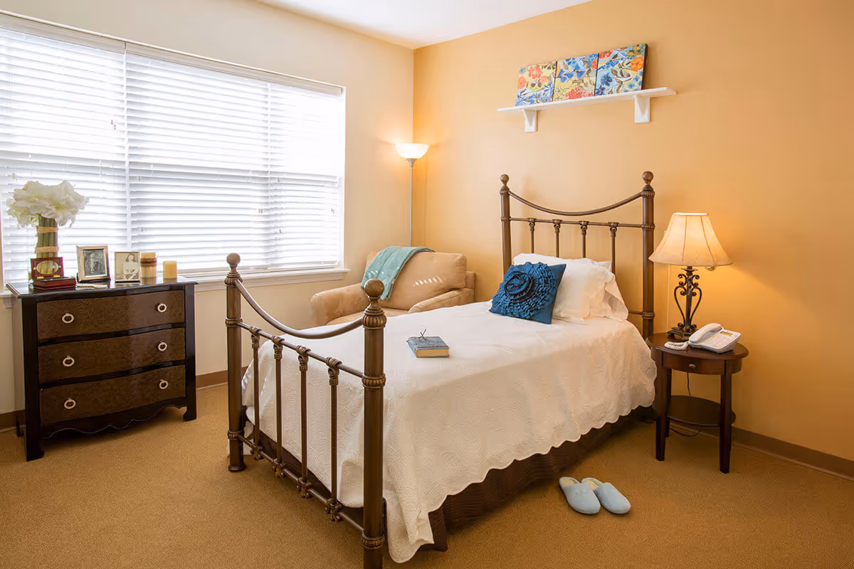 A cozy bedroom with a single metal bed frame, white bedding, and a blue decorative pillow. There is a small wooden nightstand with a lamp and a telephone, a beige armchair with a blue throw blanket, and a dark wooden dresser with framed photos and a vase of white flowers. A large window with white blinds lets in natural light, and a small shelf with colorful artwork is mounted on the wall above the bed.