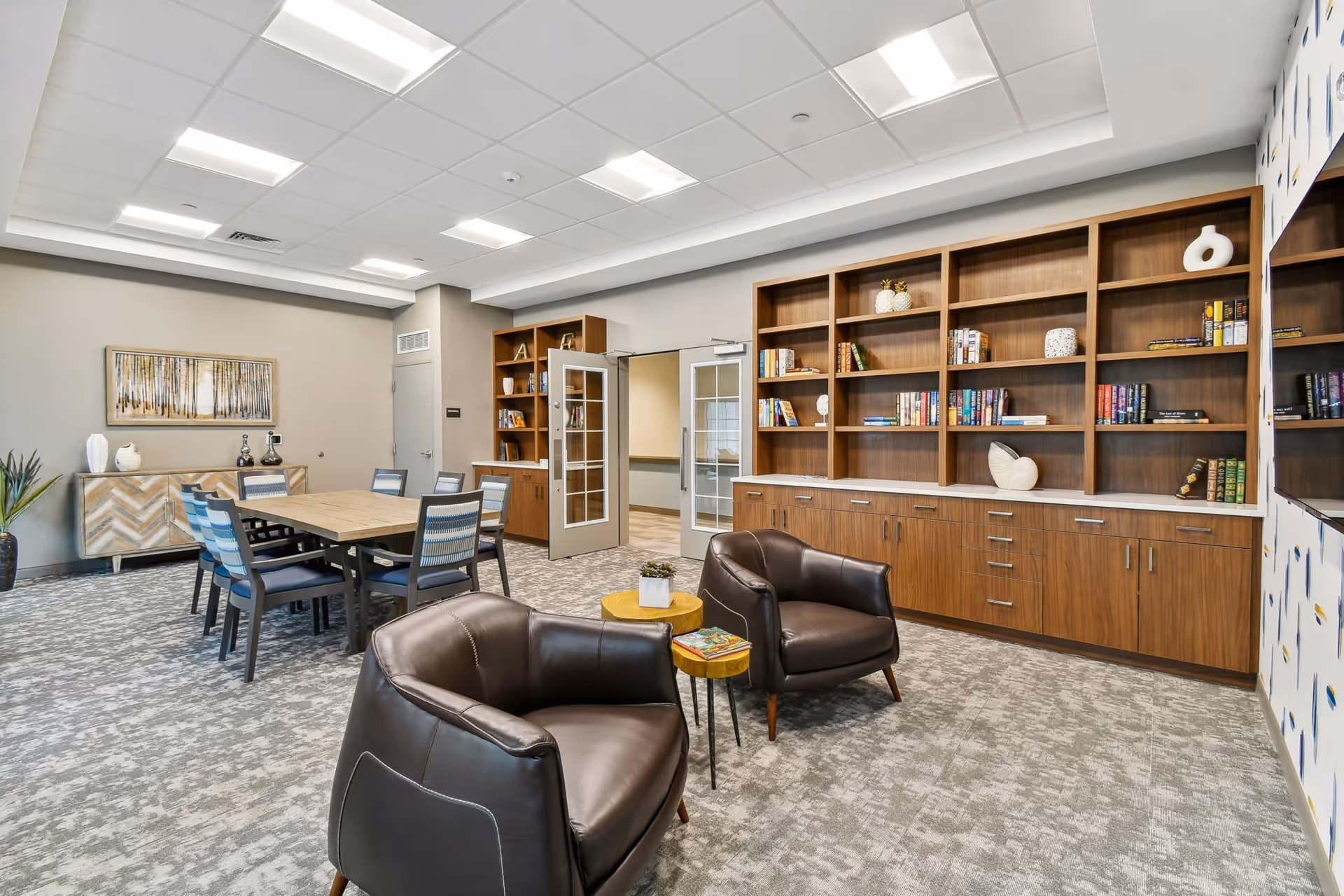 A modern senior living common area featuring two brown leather armchairs with a small wooden side table between them, a large wooden bookshelf filled with books and decorative items, a wooden dining table with eight chairs, a sideboard with decorative vases, and a framed artwork of a forest on the wall.