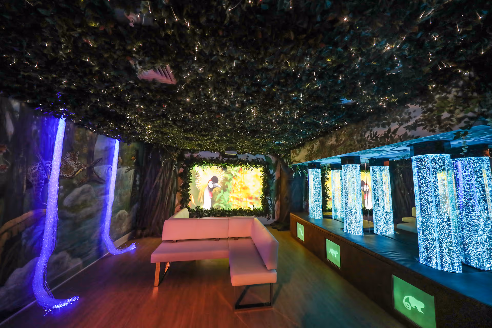 A sensory room with a nature theme featuring a ceiling covered in artificial greenery, walls painted with jungle murals, a pink L-shaped bench, illuminated bubble tubes, fiber optic lights, and a screen displaying a bird image framed by greenery.