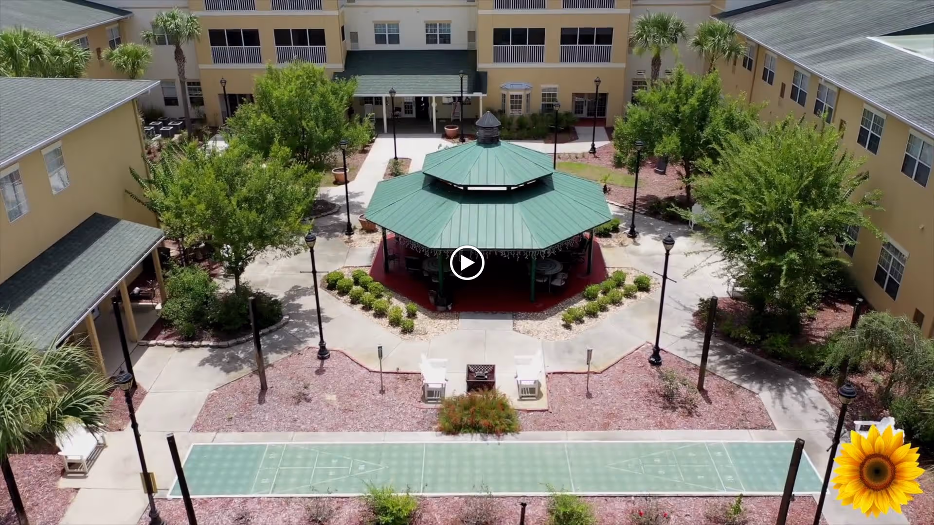 Aerial view of an outdoor courtyard area in a senior living facility with a green-roofed gazebo at the center, surrounded by trees, pathways, and buildings. There are benches and street lamps along the walkways, and a shuffleboard court is visible in the foreground.