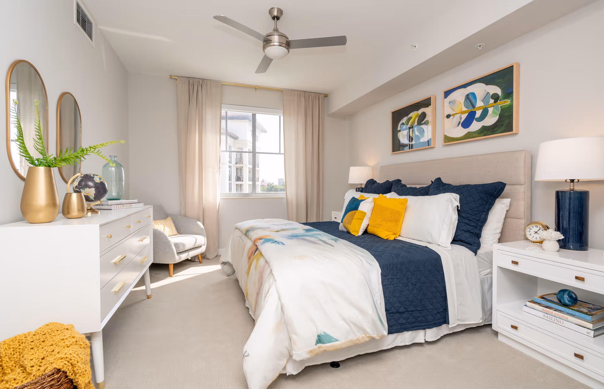 A bright and cozy bedroom with a large bed featuring navy blue and white bedding accented with yellow pillows. The room has a ceiling fan, two abstract paintings above the bed, a white dresser with decorative items including a gold vase and a globe, a comfortable gray armchair near the window with beige curtains, and a white nightstand with a blue lamp and clock.