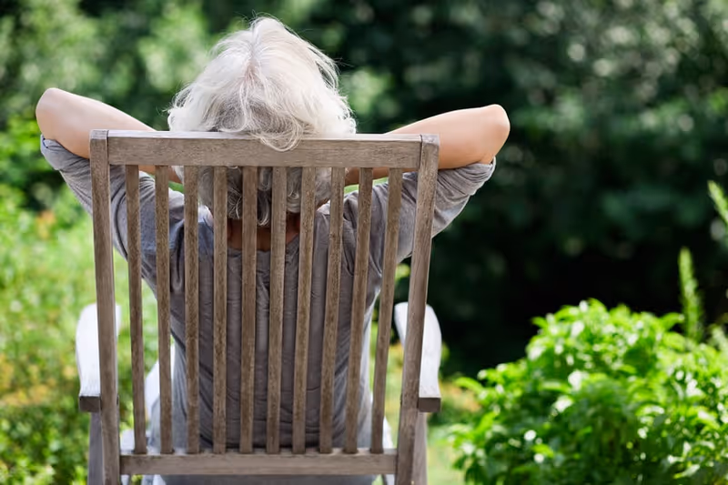 An elderly person with white hair sitting on a wooden chair outdoors, seen from behind, with arms resting on the back of the chair and surrounded by green foliage.