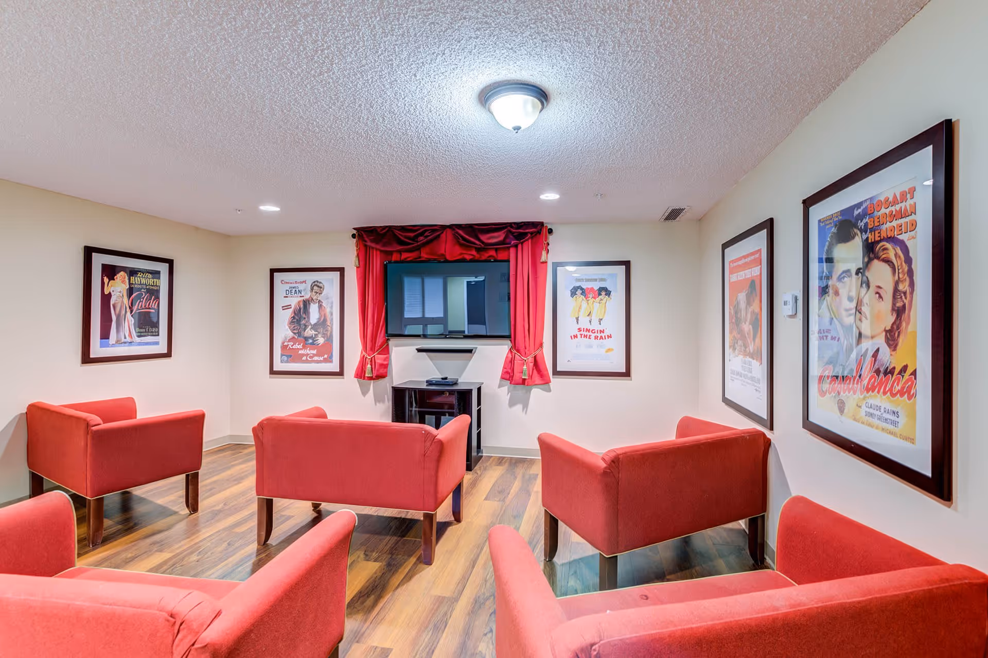 A cozy senior living common area with five red armchairs arranged facing a wall-mounted flat-screen TV. The room has wood flooring and cream-colored walls adorned with framed classic movie posters including Casablanca, Singin' in the Rain, Rebel Without a Cause, and Gilda. A red curtain decorates the window behind the TV.