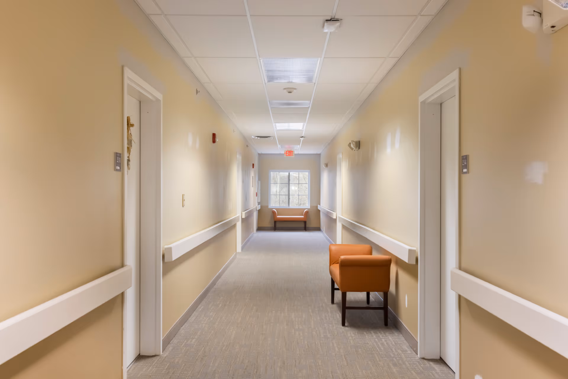 A long, well-lit hallway in a senior living facility with beige walls and carpeted floor. There are white handrails along both sides of the hallway and several closed doors with room numbers. Two orange armchairs are placed along the walls, one near the foreground on the right and one at the far end near a window. The ceiling has recessed lighting and an exit sign is visible above the window at the end of the hallway.