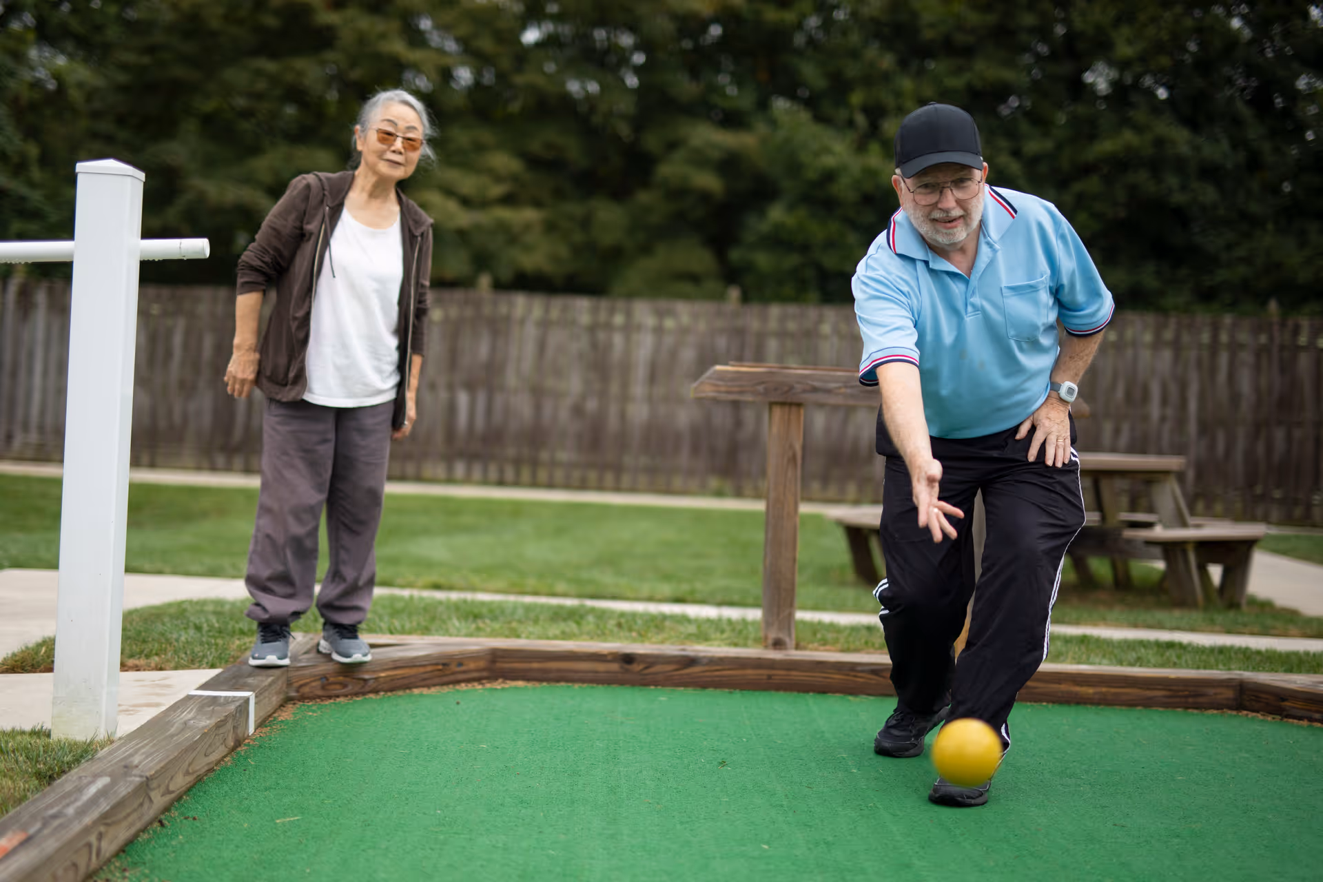 An elderly man and woman playing bocce ball outdoors on a green court. The man is in the process of rolling a yellow ball while the woman watches. They are in a fenced area with grass and trees in the background.