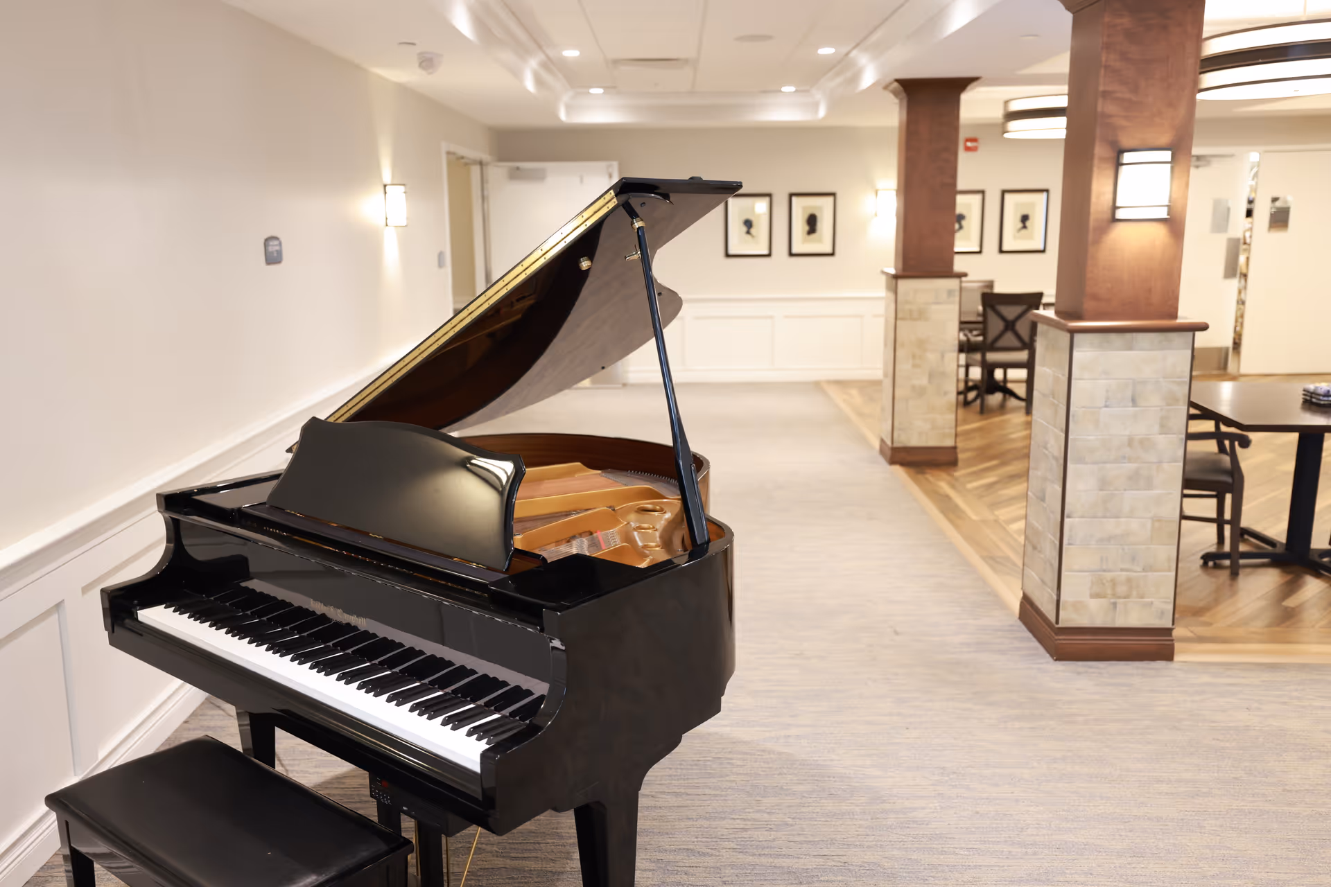 A black grand piano with its lid open is positioned in a well-lit hallway or common area of a senior living facility. The space features light-colored walls, carpeted flooring, wooden columns with stone accents, and framed silhouette artwork on the walls. There are tables and chairs visible in the background.