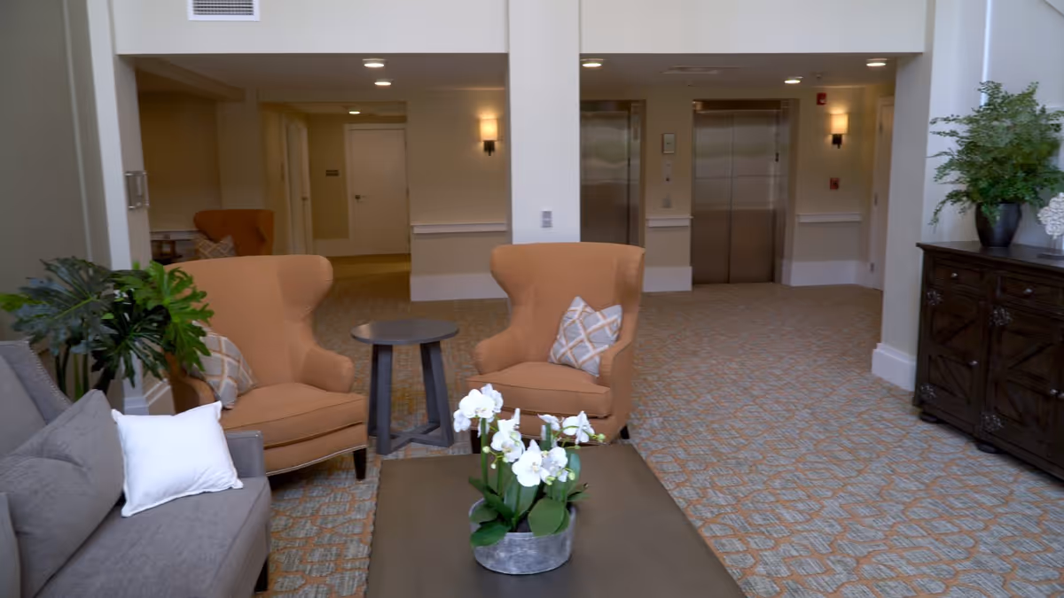 A bright facility lobby with armchairs, a coffee table with flowers, potted plants and elevators in the background.