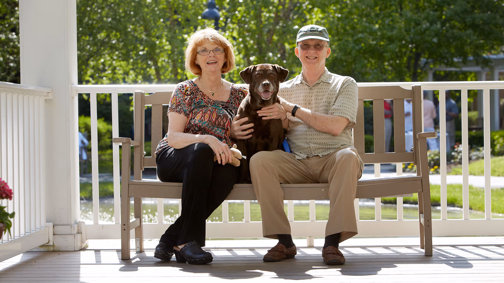 An elderly couple sitting on a bench on a porch with a brown dog between them. They are smiling and the background shows a green outdoor area with trees and a walkway.