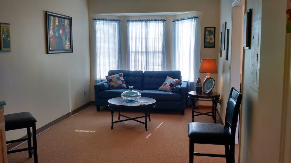 A cozy living room with a blue sofa adorned with two patterned pillows, positioned in front of three large windows with sheer white curtains. In front of the sofa is a round coffee table with a glass decorative piece. To the right of the sofa is a side table with a lamp and a framed picture. Two black chairs are placed on either side of the room. The walls are decorated with framed artwork.