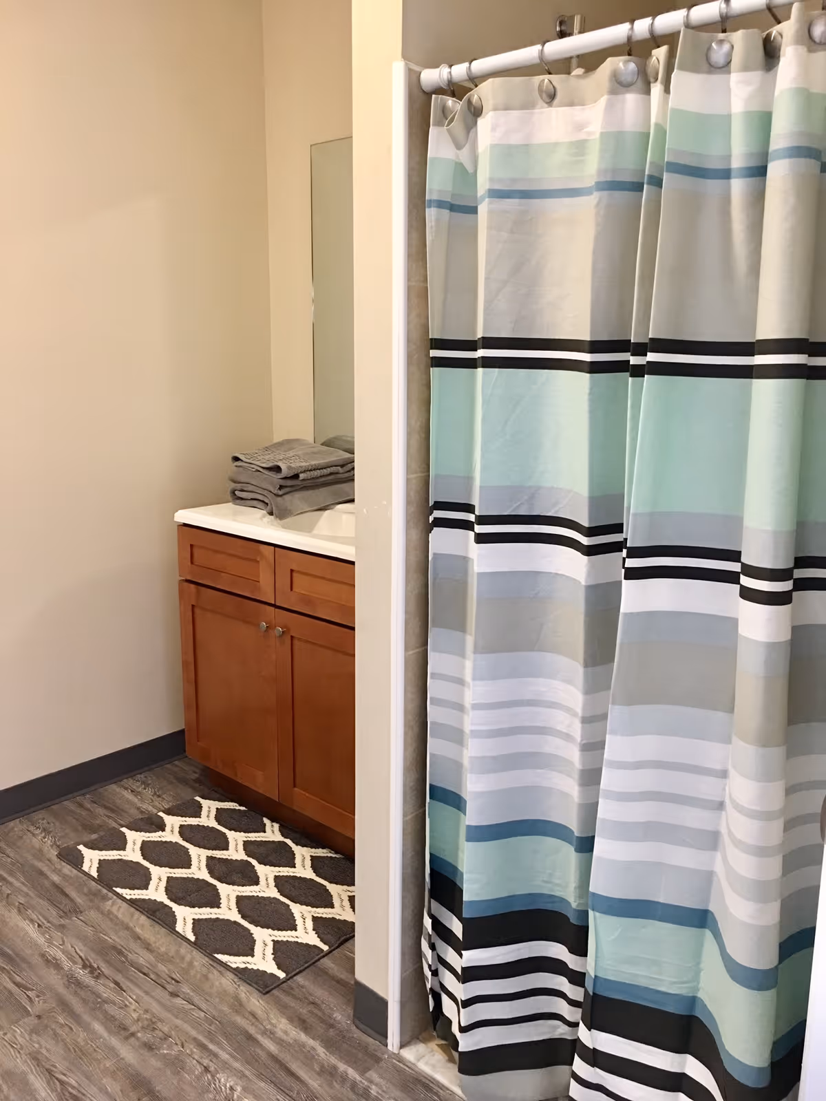 A bathroom with a wooden vanity cabinet topped with a white sink and folded gray towels. Next to the vanity is a shower area with a striped shower curtain in shades of blue, gray, black, and white. The floor has wood-like vinyl flooring and a patterned gray and white rug in front of the vanity.