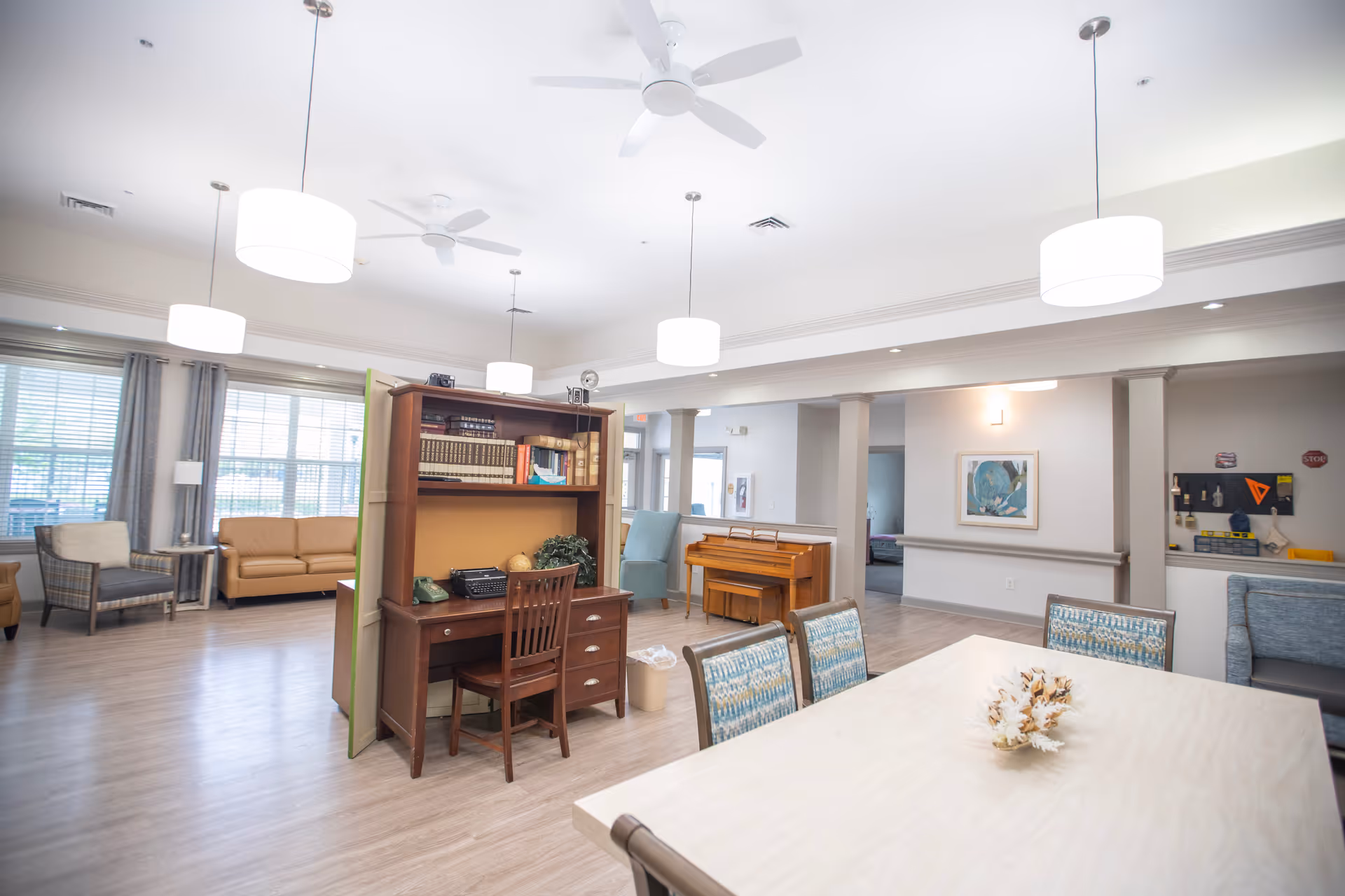 A spacious and well-lit common area in a senior living facility featuring a wooden desk with books and a chair, a piano against the wall, several seating areas with armchairs and sofas, a large table with chairs, and modern ceiling lights and fans.