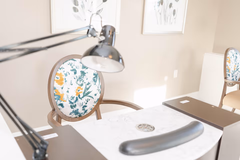 A close-up view of a manicure station with a white marble tabletop, a black adjustable desk lamp, and a floral-patterned chair with wooden arms. The background shows a beige wall with two framed botanical prints.