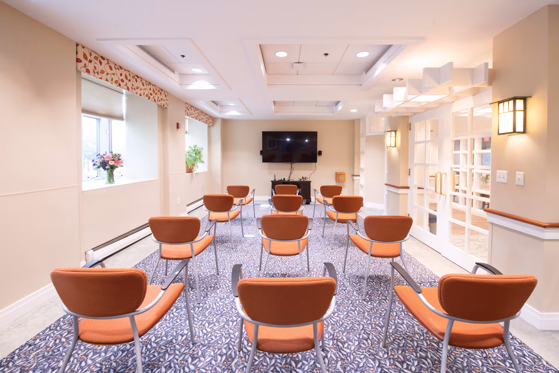 Well-lit activity room with orange chairs arranged in rows facing a wall-mounted television.