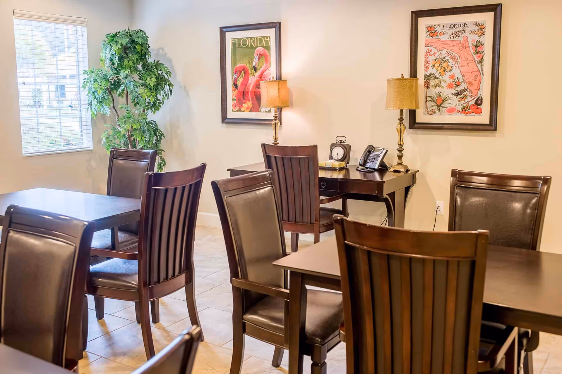 Dining room with dark wooden tables and chairs, framed Florida artwork, table lamps, a desk, and a potted plant by a window.