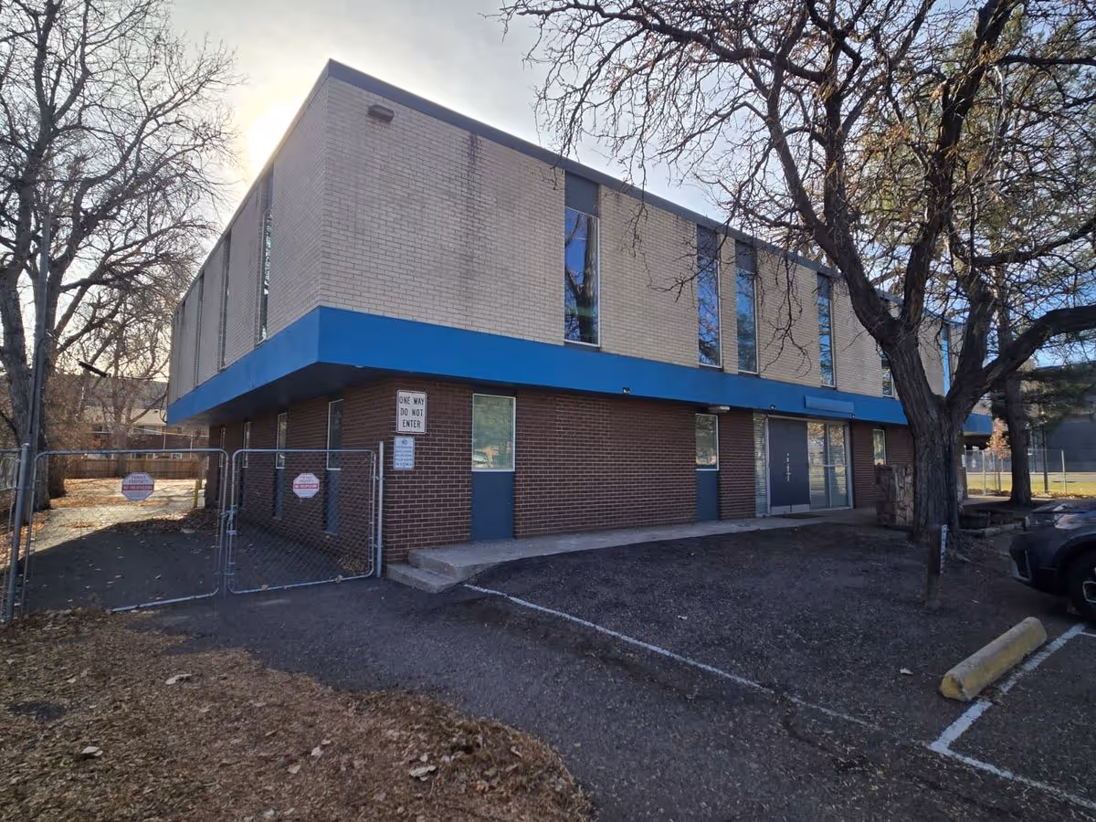 Two-story brick and beige building with blue trim, trees, a chain-link fenced entrance, and a small parking area in front.