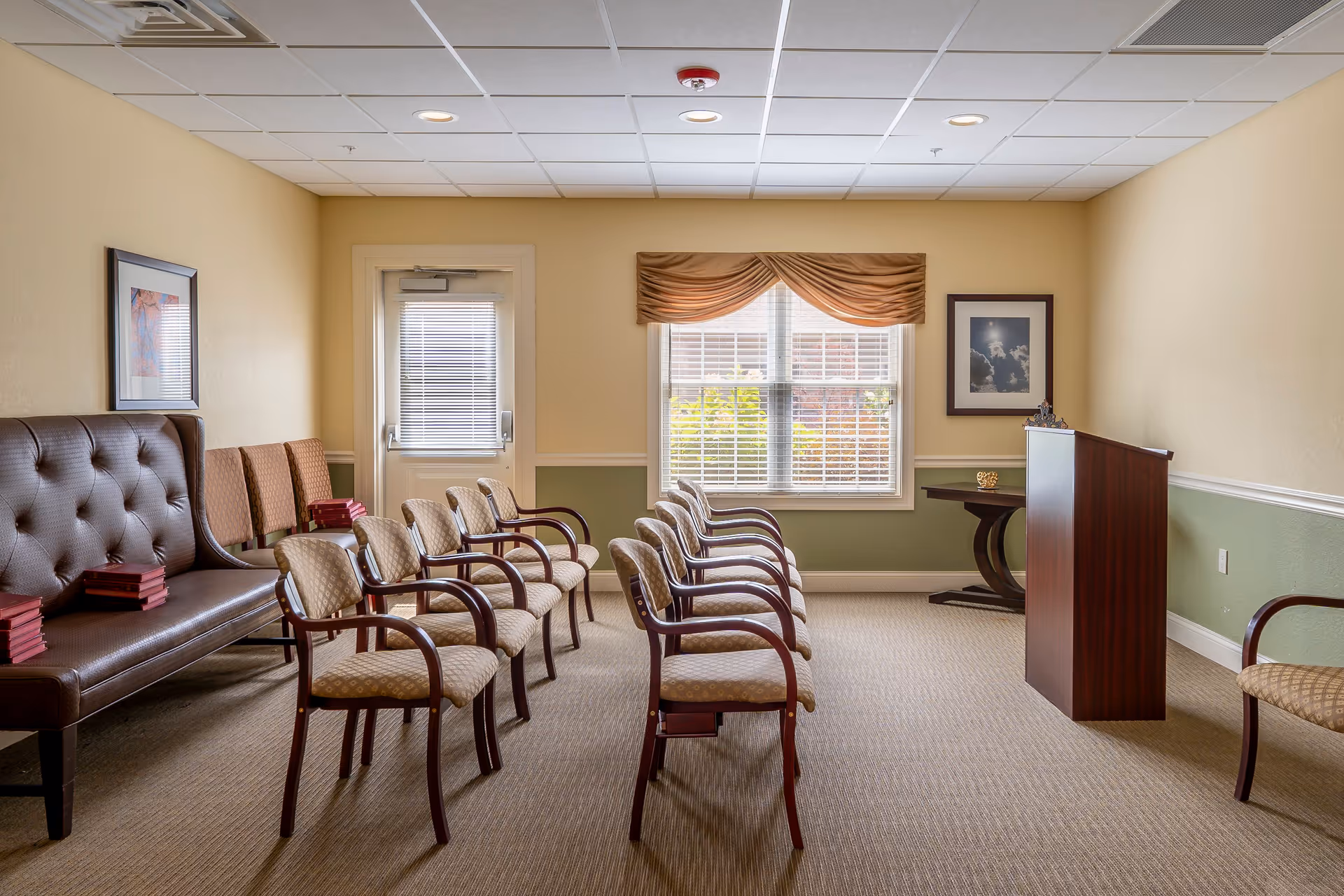 A small meeting or presentation room with two rows of beige cushioned chairs with wooden armrests facing a wooden podium. There is a brown tufted bench along the left wall with several red books stacked on it. The room has beige walls with a green wainscot, a window with blinds and a valance, a door with a window, and framed artwork on the walls.