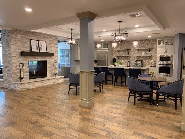 A spacious interior common area featuring a modern fireplace with stone surround and two lantern-style candle holders on the hearth. To the right, there is a kitchen area with a large island, bar stools, and hanging light fixtures. In the foreground, there is a round table with four chairs. The flooring is wood, and the space is well-lit with recessed ceiling lights.