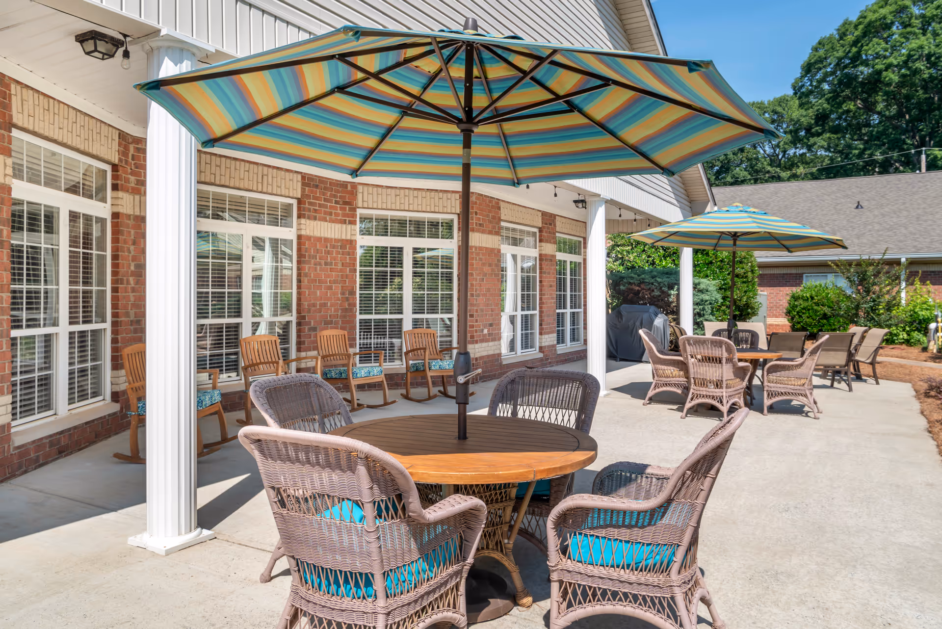 Outdoor patio area at Brookdale New Hope featuring round wooden tables with wicker chairs that have blue cushions. Each table is shaded by a large striped umbrella. The patio is adjacent to a brick building with large windows and white pillars. Additional seating includes wooden rocking chairs along the building wall. The area is surrounded by greenery and shrubs under a clear blue sky.