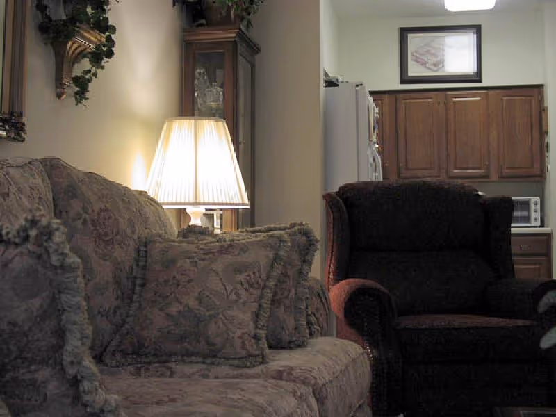 Cozy living area showing a patterned sofa with decorative pillows, a lit table lamp, an upholstered armchair, and a view into a small kitchen with wooden cabinets.