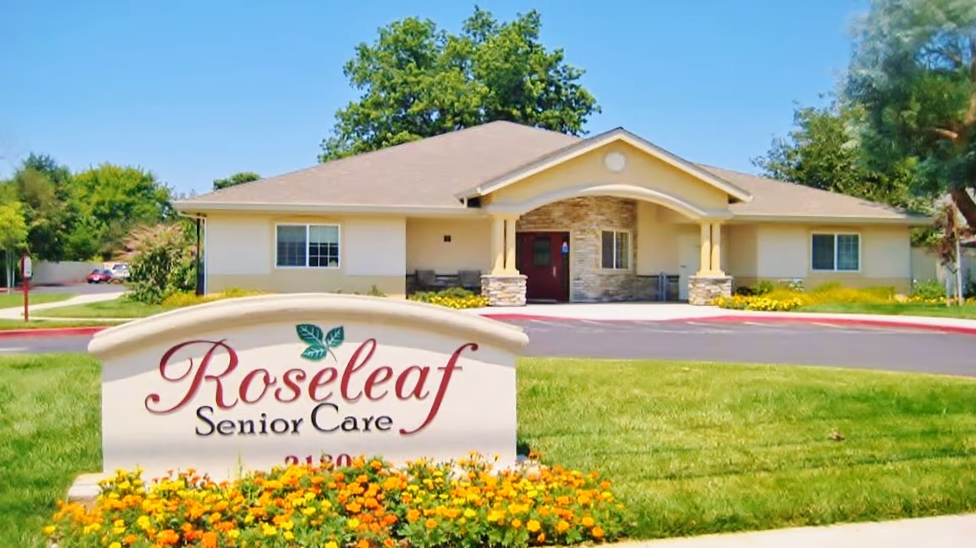 Front exterior view of Roseleaf Senior Care facility with a single-story building, a manicured lawn, and a sign in the foreground surrounded by yellow and orange flowers under a clear blue sky.