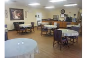 A nursing home dining room with several round tables covered with white tablecloths and wooden chairs arranged around them. The room has light-colored walls, a wooden floor, and fluorescent ceiling lights. There is a framed picture on the wall and a clock above a counter area with coffee machines and other appliances.
