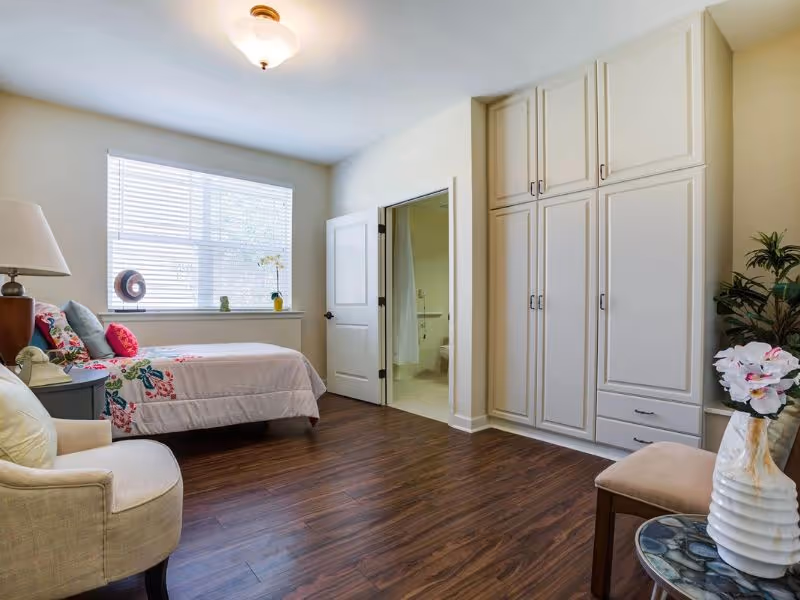 A bright and clean bedroom with a single bed covered in a white quilt with colorful floral patterns. There is a beige armchair, a small round table with a white vase holding flowers, and a large window letting in natural light. A door leads to an adjacent bathroom, and there are built-in white cabinets along one wall. The floor is wooden.