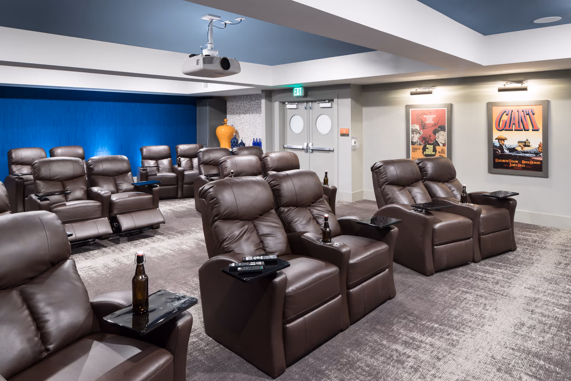 A cozy theater room with multiple rows of brown leather recliner chairs, each equipped with a small black side table holding remote controls and bottles. The room has a blue accent wall, a ceiling-mounted projector, and two framed vintage movie posters on the wall near double doors.