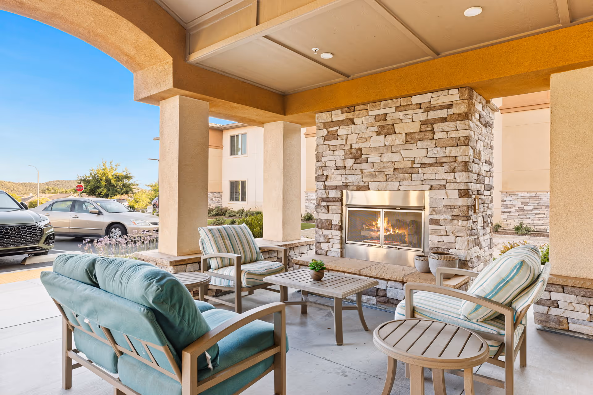 Covered outdoor seating area with cushioned chairs and a stone fireplace near the facility entrance.