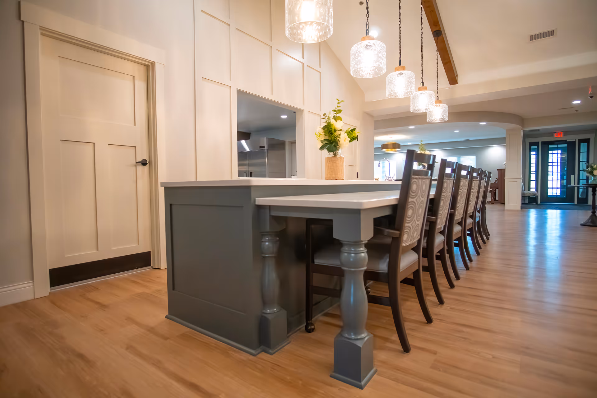 Open communal dining area with a long island table, multiple chairs, pendant lights, and wood flooring.