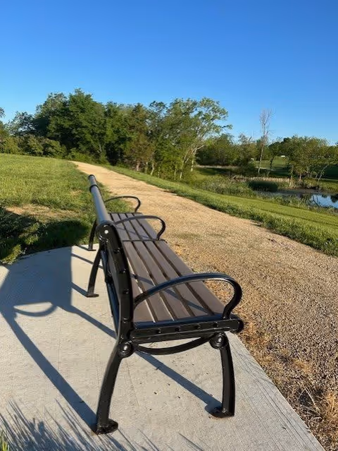 A metal and wood bench on a concrete pad next to a gravel walking path in a green park-like area with trees and a small pond under a clear blue sky.