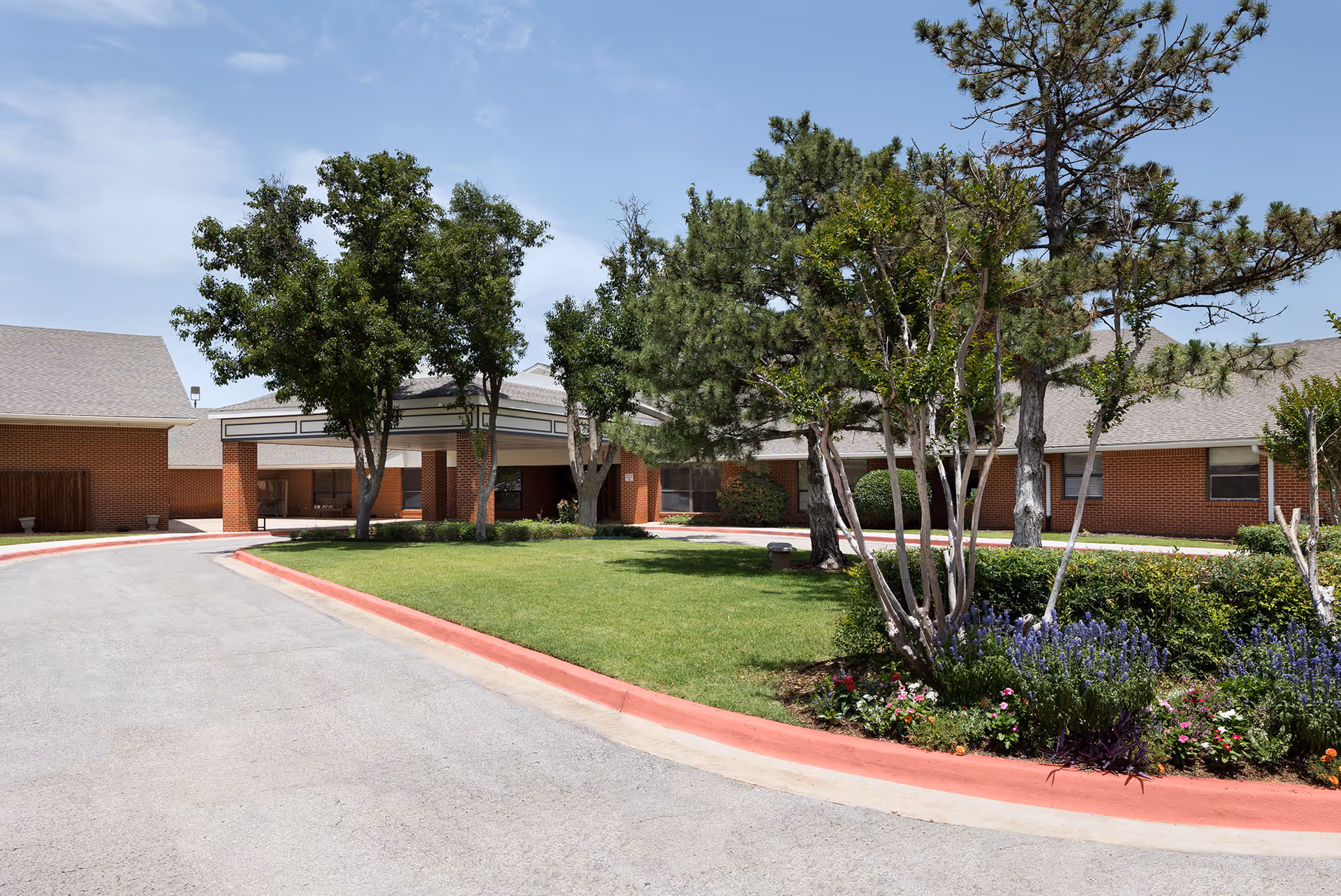 Front exterior of a single-story brick skilled nursing facility with a covered entrance, circular driveway, lawn, trees, and flower beds.
