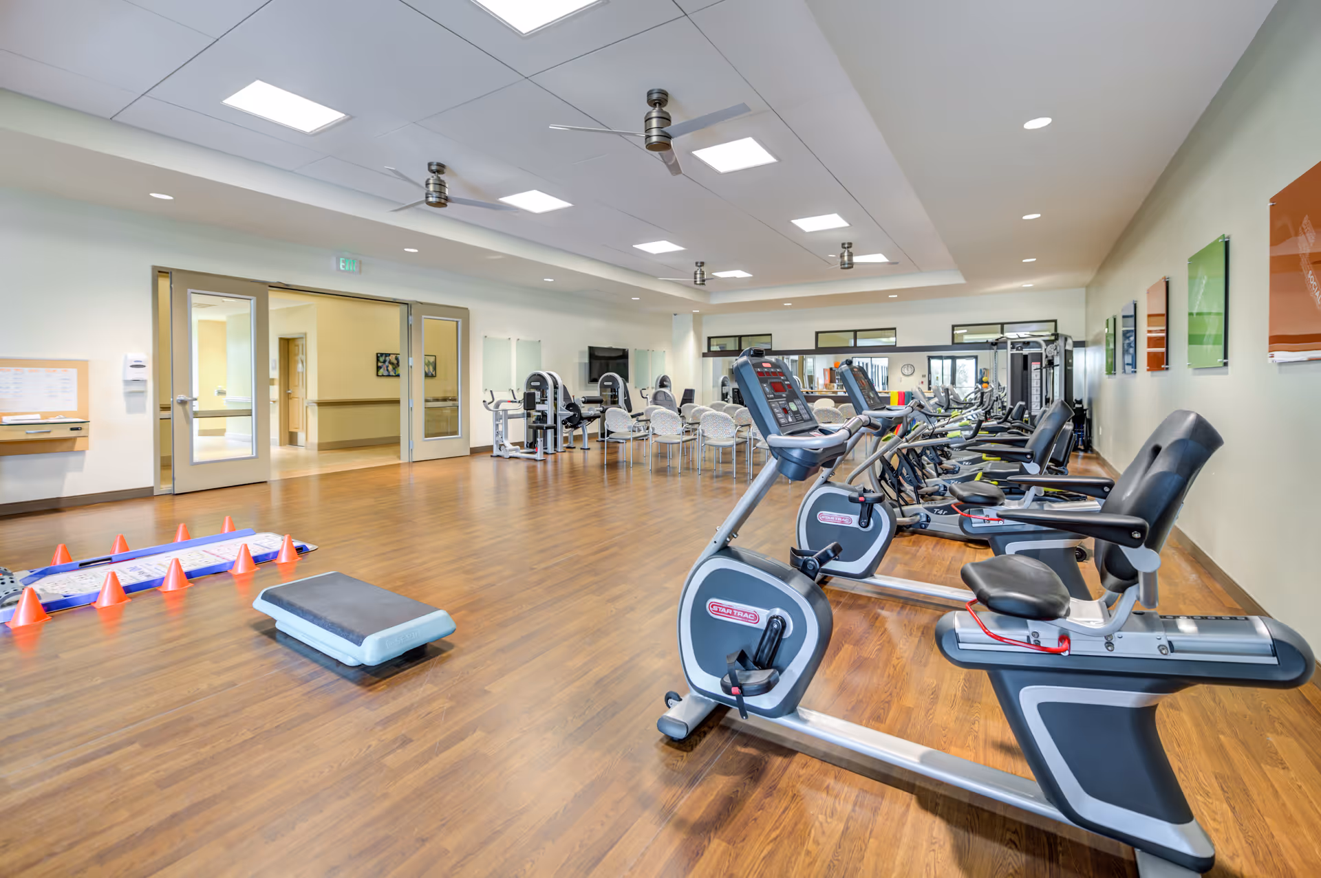 A spacious fitness room in a senior living facility with exercise equipment including stationary bikes, treadmills, and weight machines. The room has wooden flooring, bright overhead lighting, ceiling fans, and colorful wall art. There are also chairs and tables in the background near glass doors leading to a hallway.