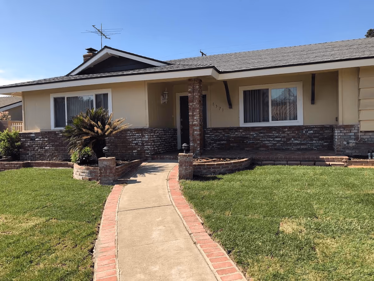 Single-story ranch-style house front with a brick facade, central walkway and a green lawn.