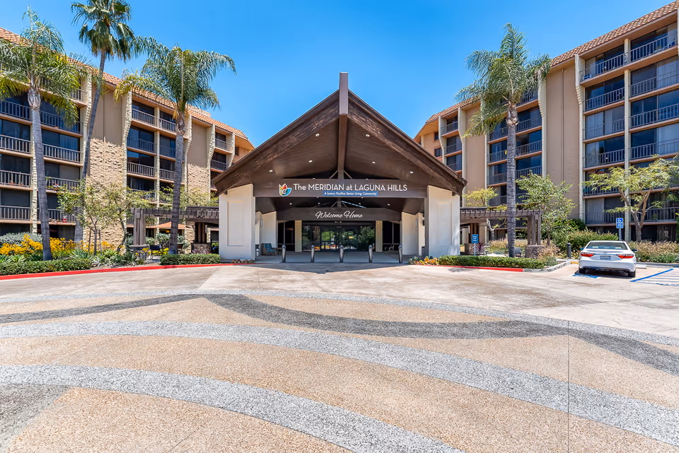 Front entrance of The Meridian at Laguna Hills senior living community with a covered porte-cochere, flanked by multi-story buildings and palm trees.