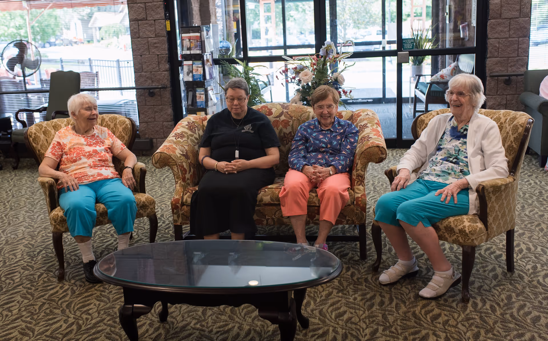 Four elderly women sitting on upholstered chairs and a loveseat in a well-lit common area with large windows and a glass coffee table in front of them. The room has patterned carpet and a floral arrangement on the loveseat.