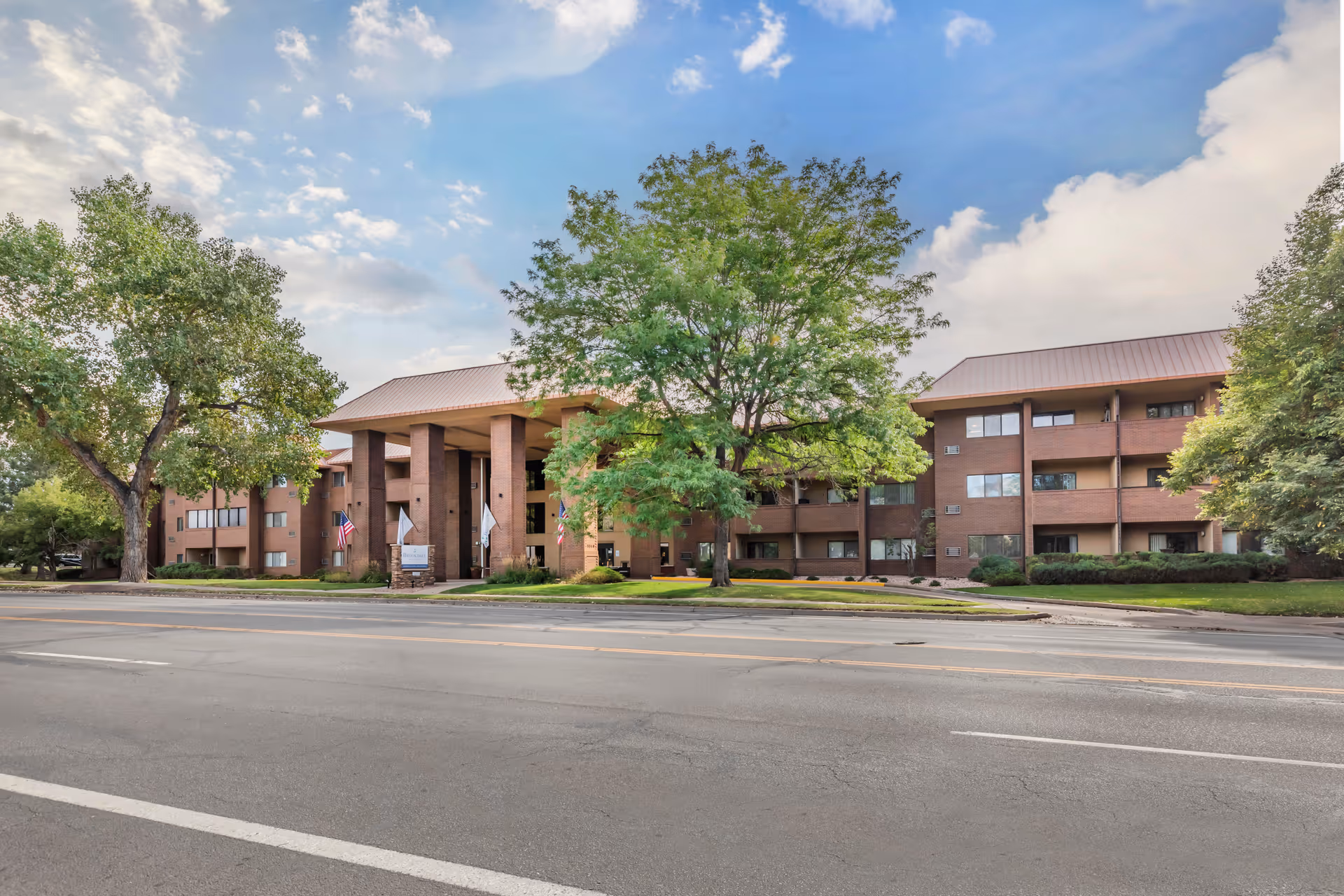 Exterior view of a three-story brick building with a covered entrance supported by large columns, surrounded by trees and greenery under a partly cloudy sky.