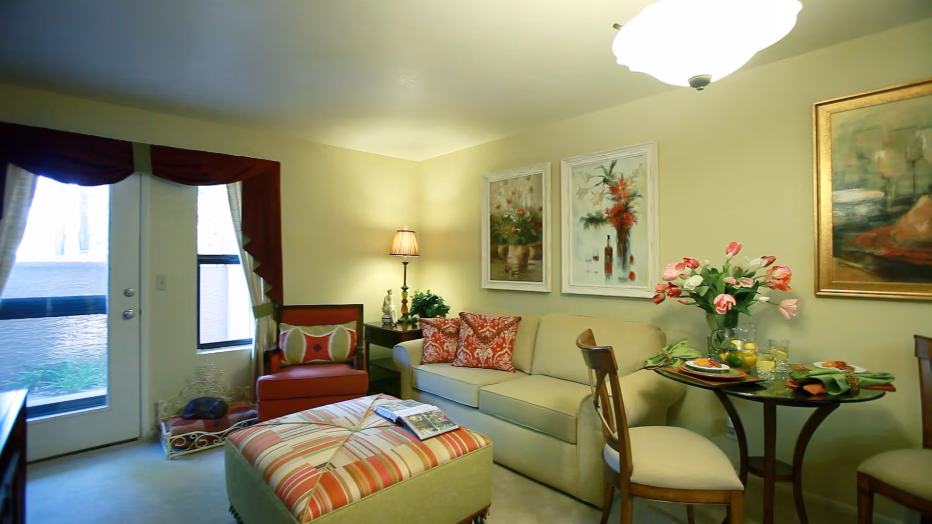 A cozy living room area with a beige sofa adorned with red and patterned cushions, a striped ottoman with a magazine on top, a red armchair with a decorative pillow, a small side table with a lamp and plant, and a round dining table set with plates, glasses, and a vase of pink tulips. The room has light yellow walls decorated with framed floral paintings and a door with a window covered by red curtains.