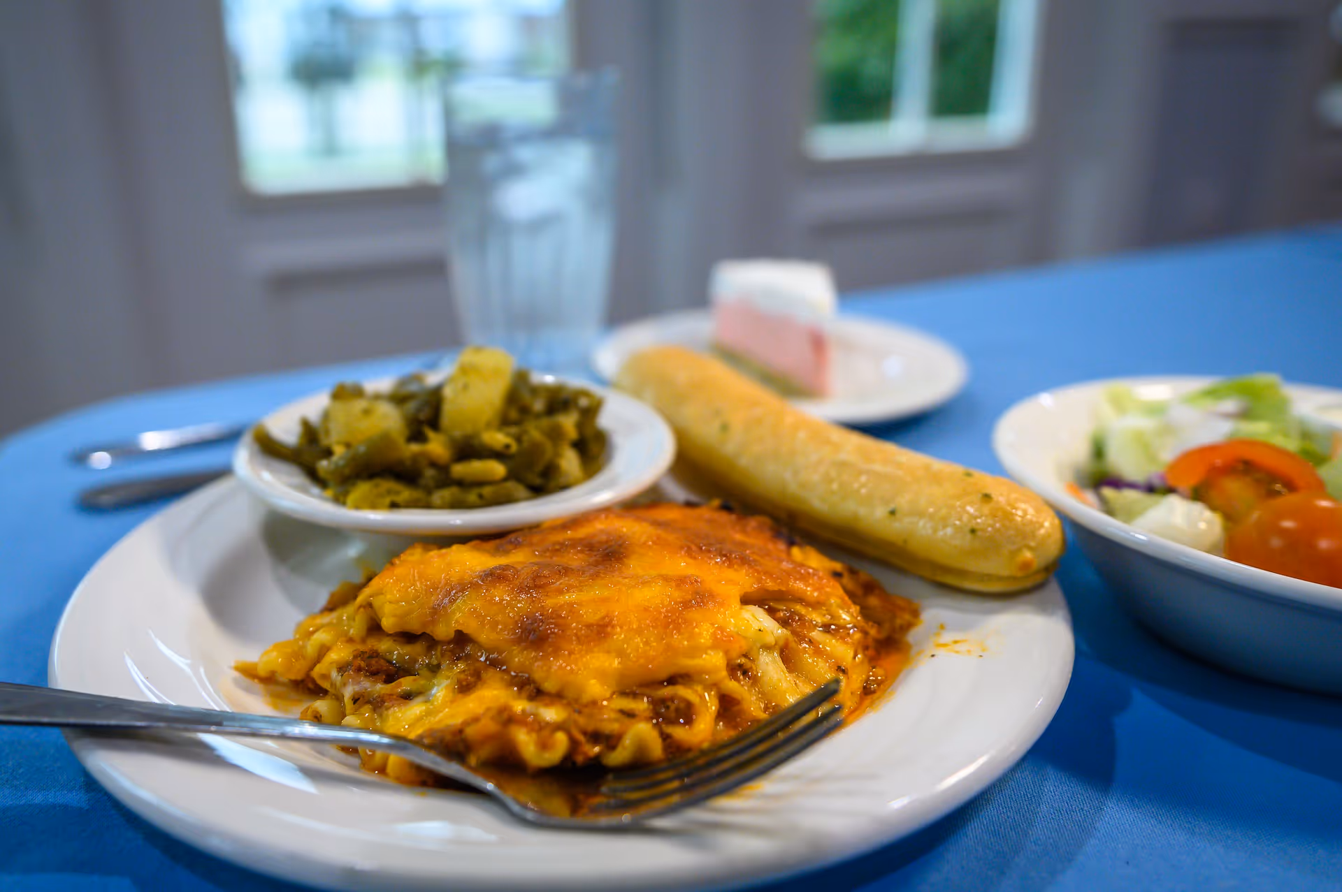 A plate of food on a blue tablecloth featuring a serving of lasagna with melted cheese, a breadstick, and a bowl of cooked green beans. In the background, there is a glass of water, a small slice of cake on a plate, and a bowl of salad with cherry tomatoes and lettuce.