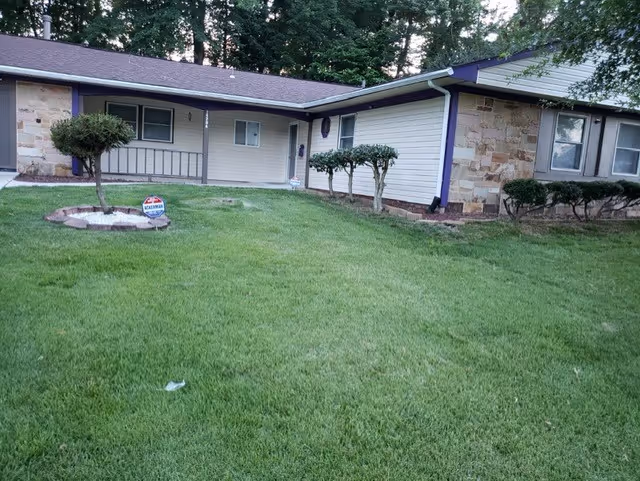 Front exterior view of a single-story residential building with stone and siding walls, a well-maintained green lawn, and trimmed bushes in front of the entrance.