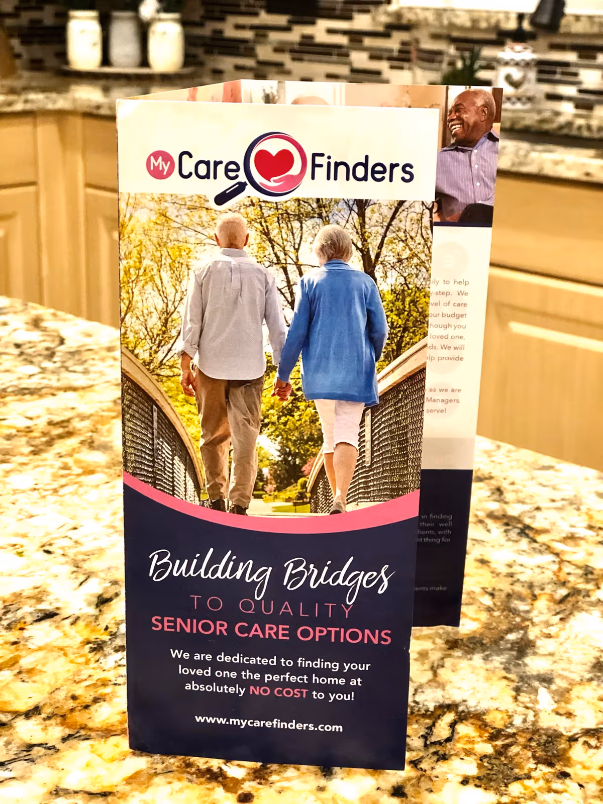 A brochure for My Care Finders standing on a granite countertop in a kitchen. The brochure features an image of an elderly couple walking hand in hand on a bridge surrounded by trees. The text on the brochure reads: 'Building Bridges to Quality Senior Care Options. We are dedicated to finding your loved one the perfect home at absolutely NO COST to you! www.mycarefinders.com'.