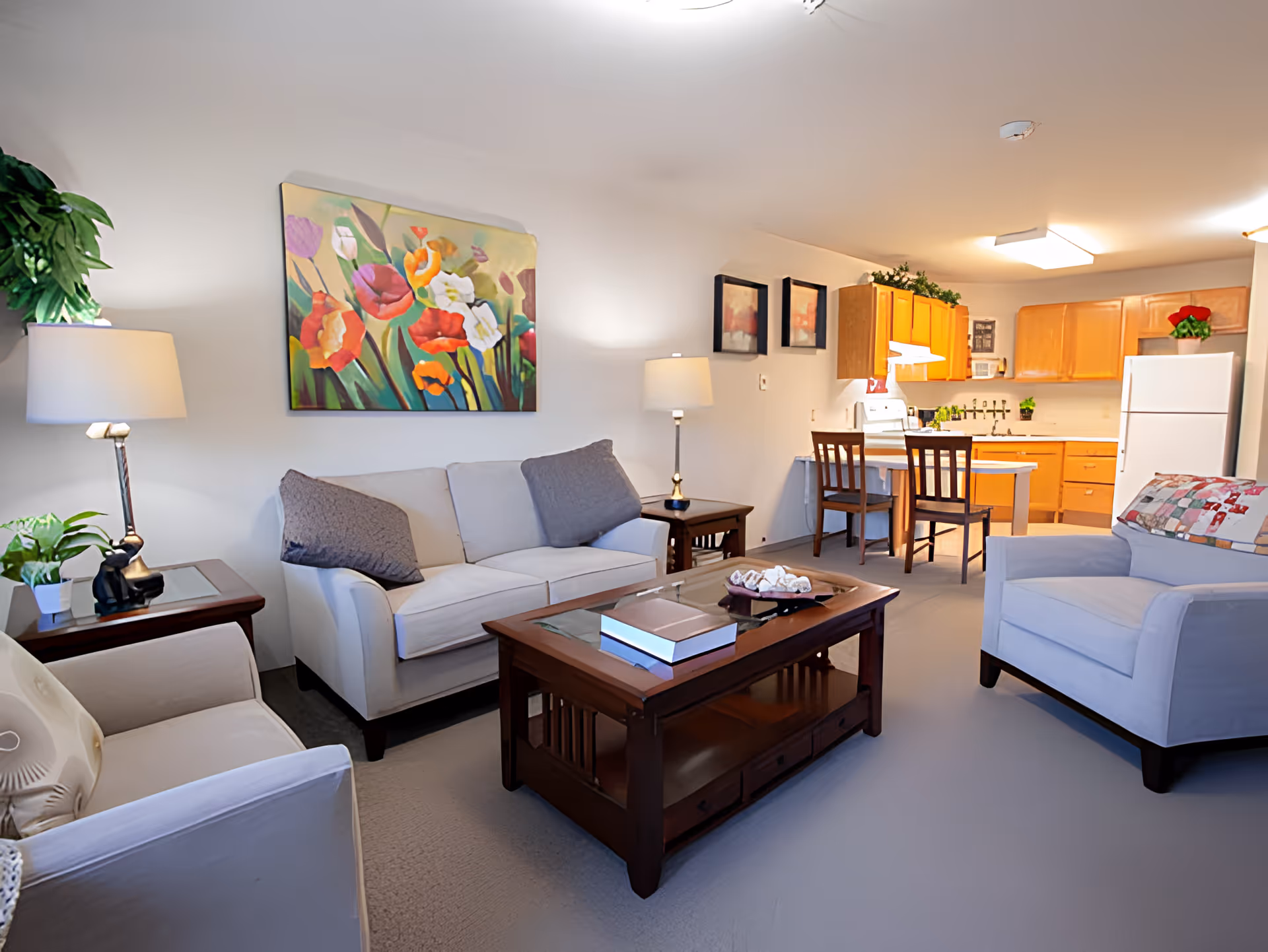 A cozy living room area with beige sofas and armchairs, a wooden coffee table with books and decorative items, two side tables with lamps and plants, colorful floral artwork on the wall, and an open kitchen with wooden cabinets, a white refrigerator, stove, and a small dining table with two chairs.