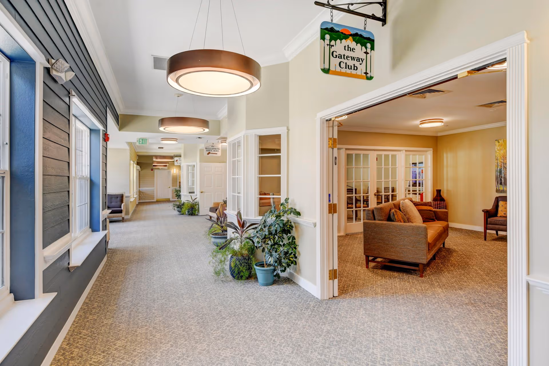 A bright and spacious hallway in a senior living facility with large windows on the left, modern circular ceiling lights, and several potted plants along the right wall. To the right, an open doorway leads to a cozy sitting area with a sofa, armchairs, and a painting on the wall. Signs hanging from the ceiling indicate 'the Gateway Club' and 'the Wellspring Town Hall.'