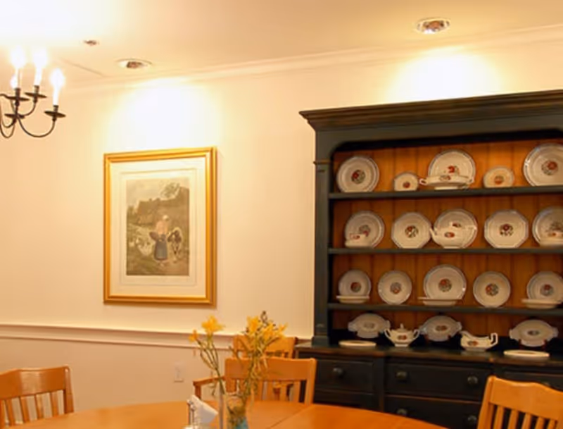 A dining room with a wooden table and chairs, a black hutch displaying decorative plates, a framed painting on the wall, and a chandelier with lit candles.