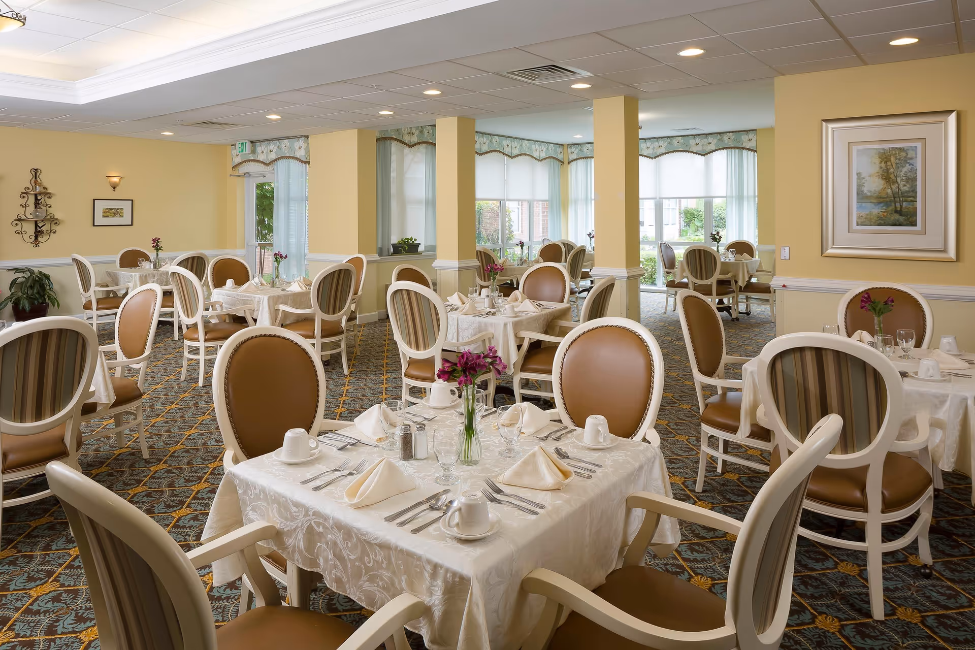 A bright and elegant dining room with multiple round and square tables covered with white tablecloths. Each table is set with folded napkins, cups, glasses, and silverware. The chairs have white frames with brown and striped upholstery. The room has large windows with sheer curtains allowing natural light to fill the space. The walls are painted yellow with white wainscoting and decorated with framed artwork and a wall sconce.