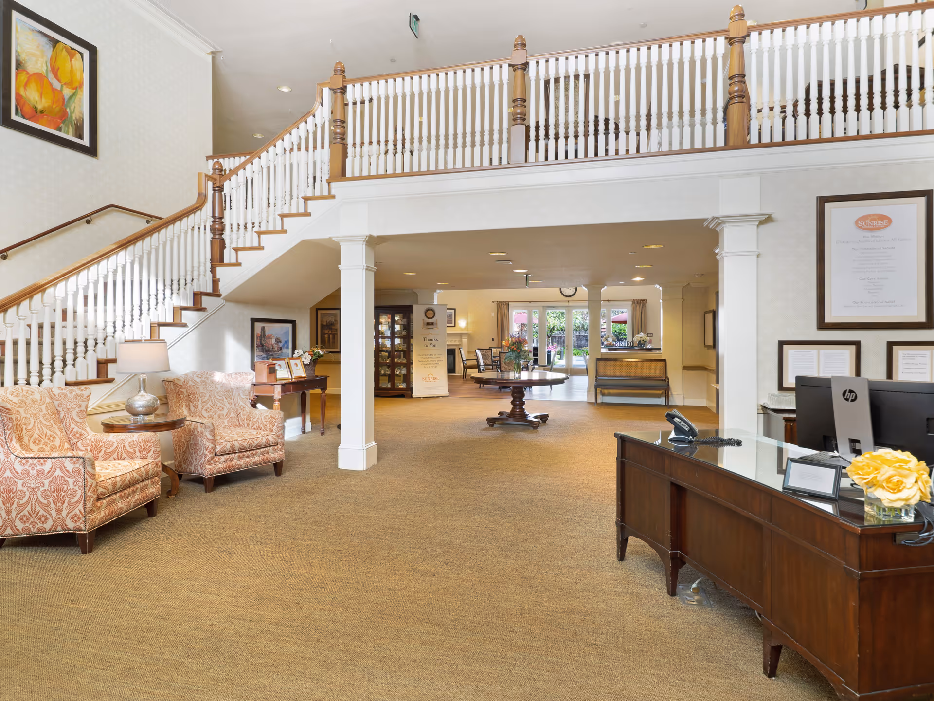 Interior view of a senior living facility lobby with a wooden reception desk on the right, two patterned armchairs with a small round table and lamp on the left, a staircase with wooden banisters leading to an upper floor, and a round table with a flower arrangement in the center. The background shows a seating area with chairs and large windows letting in natural light.