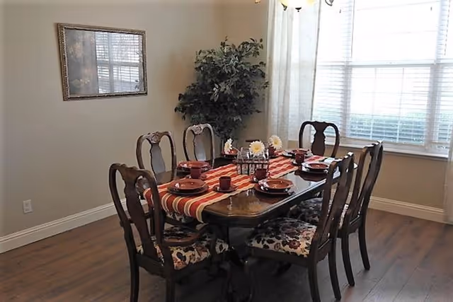 A dining room with a wooden dining table set for six people. The table has a red and white striped runner, red plates, cups, and a centerpiece with white flowers. There are six wooden chairs with floral-patterned cushions around the table. A large window with white blinds and sheer curtains lets in natural light. A framed picture hangs on the wall and a green potted plant is in the corner.