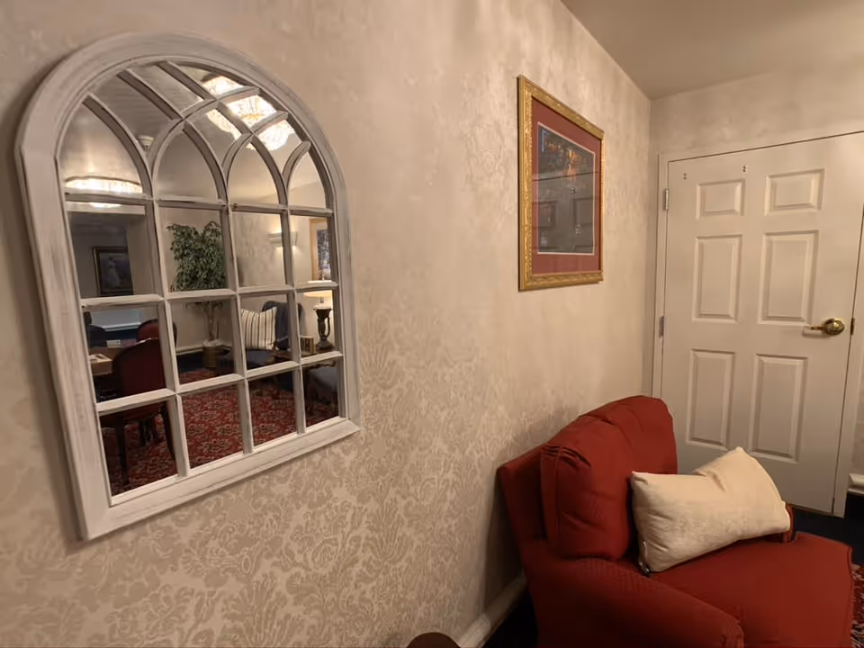 A cozy corner of an interior room with beige patterned wallpaper, a red cushioned chair with a white pillow, a white door with a brass handle, and a decorative framed picture on the wall. A large arched mirror with multiple panes reflects part of the room, showing a plant, a table, and chairs with a red carpet.