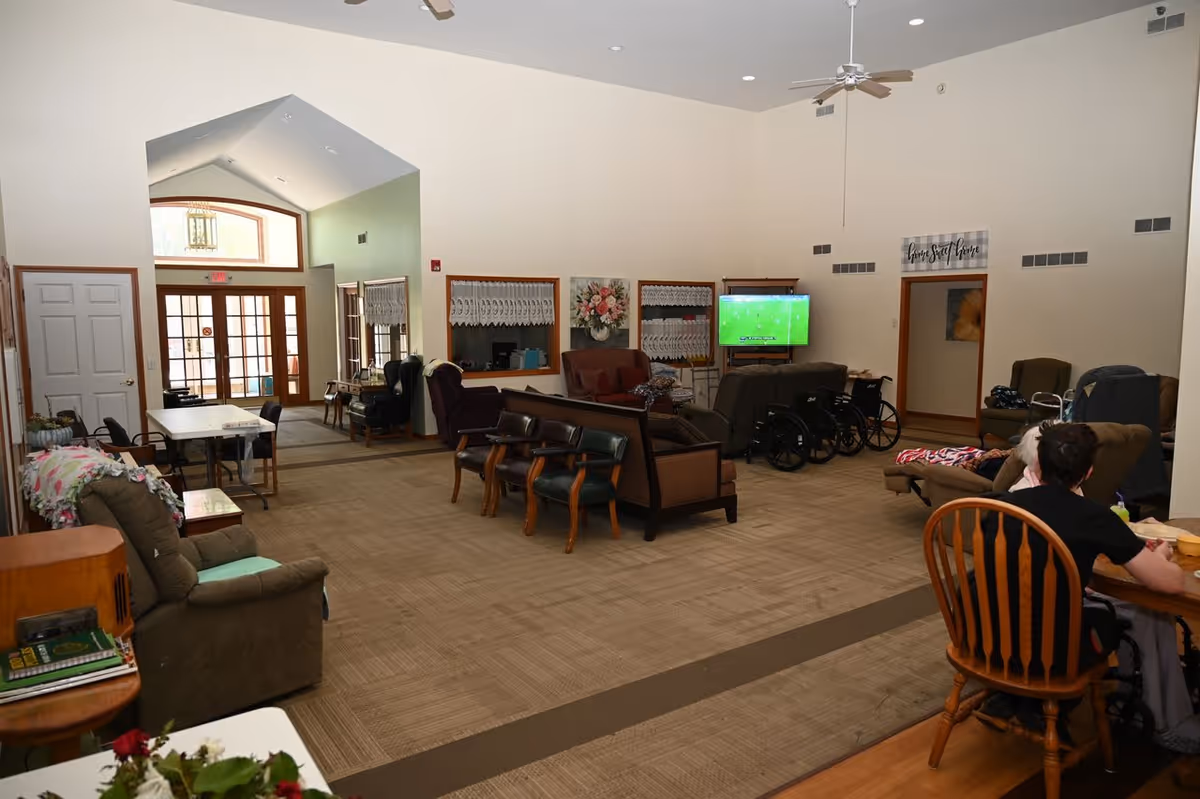 A spacious senior living common area with multiple seating options including armchairs, sofas, and wooden chairs. A television mounted on the wall is showing a sports game. There are wheelchairs lined up near the seating area. The room has high ceilings with ceiling fans, large windows with lace curtains, and a door leading to another room. Two people are seated at a wooden table on the right side of the image.