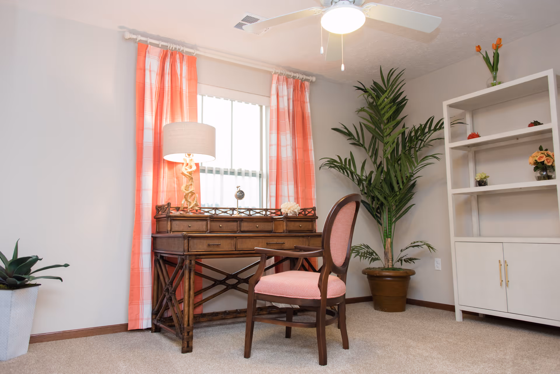 A cozy interior room featuring a wooden desk with multiple drawers, a table lamp with a beige shade, and a small clock on top. In front of the desk is a wooden chair with a pink cushioned seat and backrest. The window behind the desk is adorned with pink and white checkered curtains. To the right, there is a tall green potted plant and a white shelving unit with decorative flowers. The room has beige carpet flooring and a ceiling fan with a light.